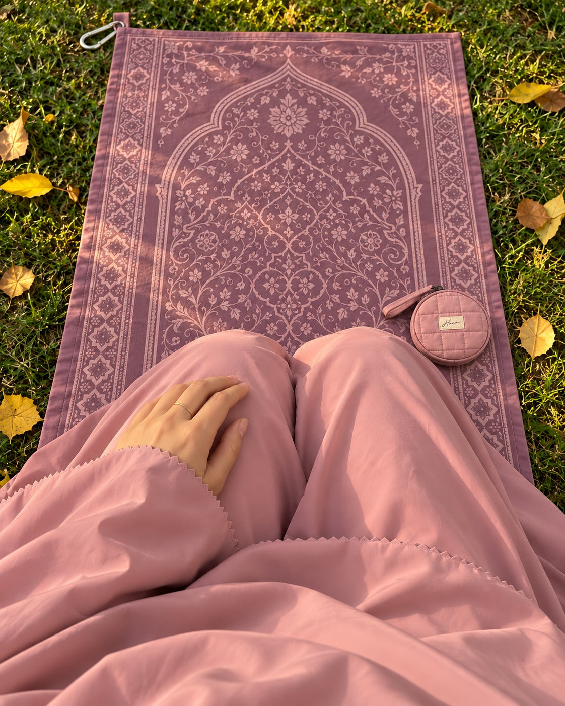 Seen from above: her lap in the Shafaq outfit on a plum travel prayer mat in a park, the closed pouch beside her knee