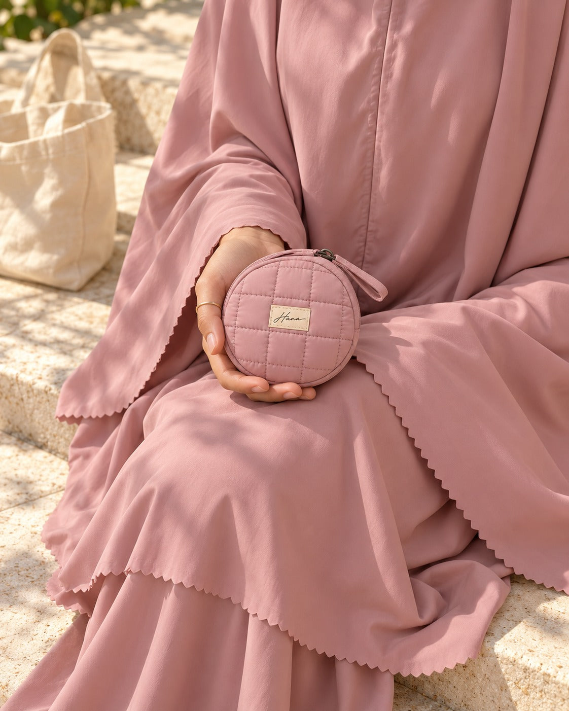 The round Hana pouch resting in her palm, the scalloped hem of the Shafaq outfit around it